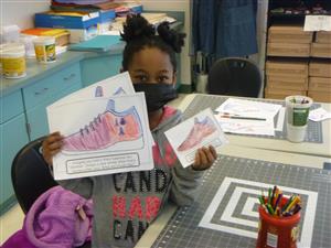 Photo of a student holding up a paper with a colorful picture of a school she designed.